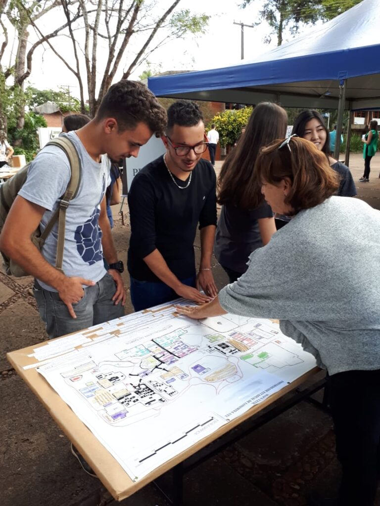 DESCRIÇÃO DA IMAGEM: A pedagoga Eliane Guimarães apresentando um mapa tátil da UEL para 2 estudantes com deficiência visual durante atividade na Feira de Profissões da UEL/ 2019, ao fundo há alunos sob uma tenda.
