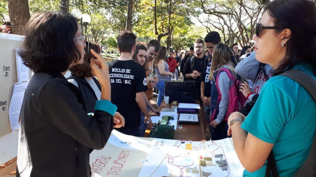 DESCRIÇÃO DA IMAGEM: Em primeiro plano a Profa. Karen conversa em LIBRAS com um estudante surdo durante atividade na Feira de Profissões 2019, ao fundo há vários estudantes em pé em volta da mesa.